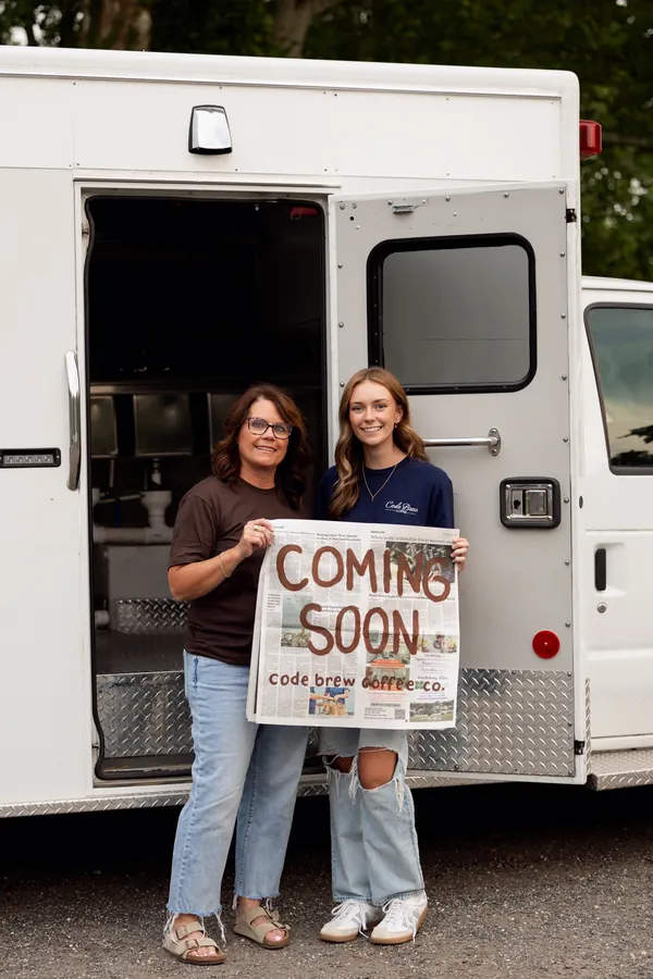 Deb and Abbie in front of the open ambulance, holding a hand-painted Coming Soon, Code Brew Coffee Co. sign