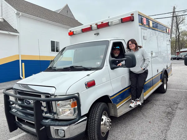Rick in the driver’s seat and Deb standing on the step of the white ambulance with blue and gold stripes, the day they got it